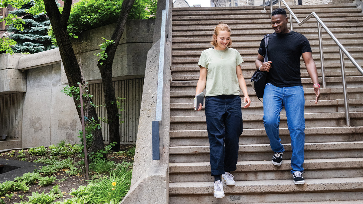 Deux étudiants descendent un escalier extérieur sur un campus, en discutant, entourés de végétation.